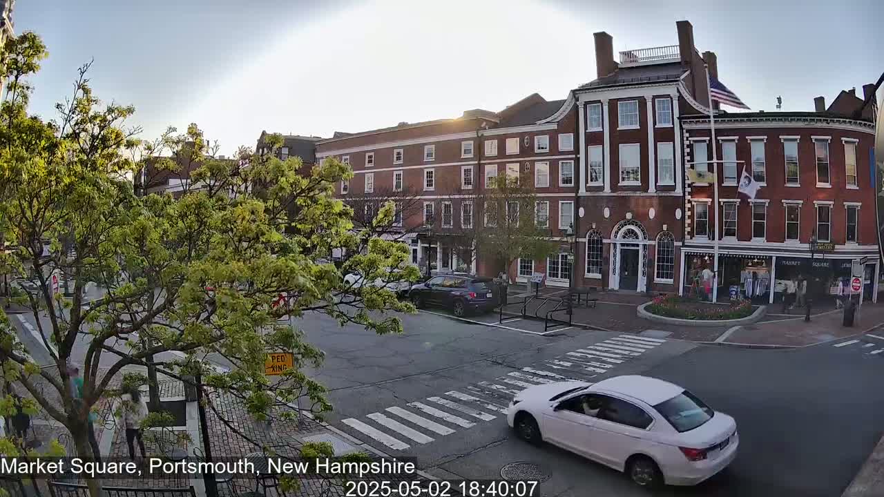 A white car drives through a tree-lined town square past brick buildings under a mostly clear evening sky.