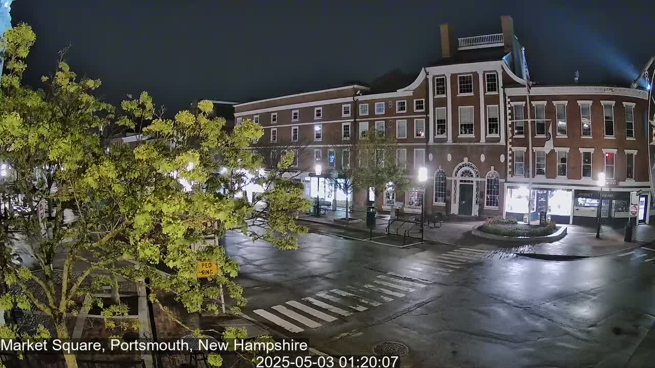 A wet, nighttime street scene shows a brick building with illuminated windows and a few trees with green leaves in a town square.