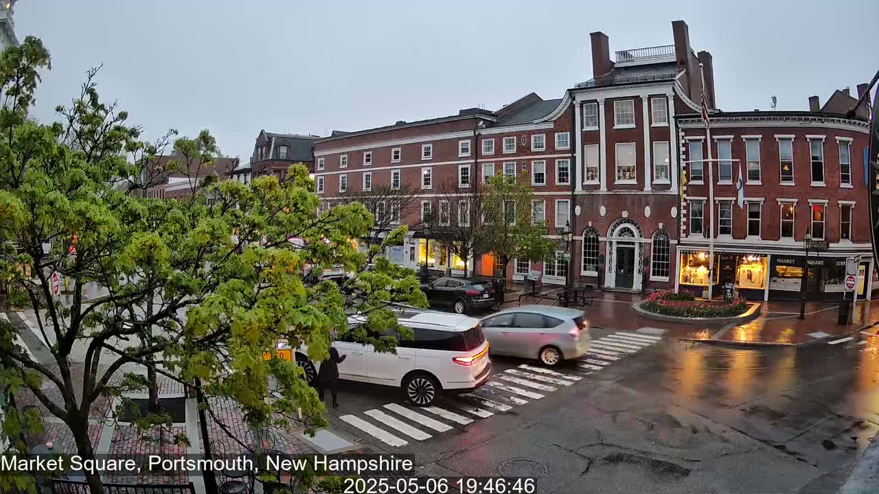 A wet, overcast town square features several brick buildings, lush green trees, and a few cars at a crosswalk.