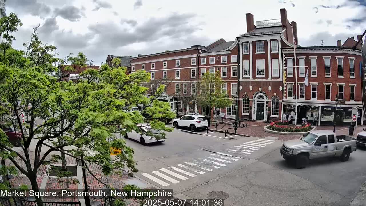 A partly cloudy day shows a brick building and several cars in a town square, with lush green trees lining the streets.