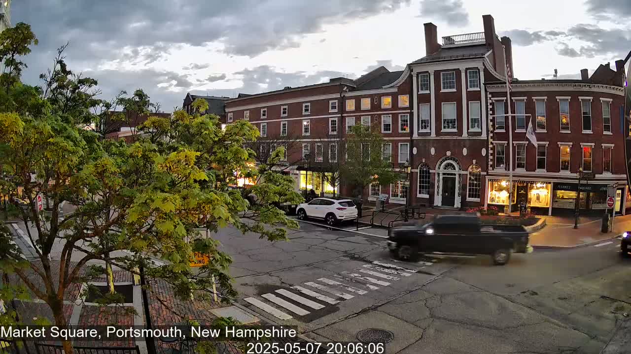 A mostly cloudy evening scene shows a brick-building-lined town square with cars driving on the street, and lush green trees in the foreground.