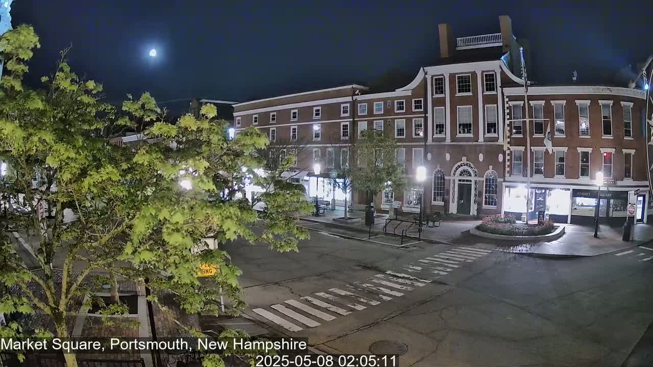 A nighttime view of a brick building in a town square under a mostly clear sky with a visible moon, illuminated by streetlights, with leafy green trees in the foreground.