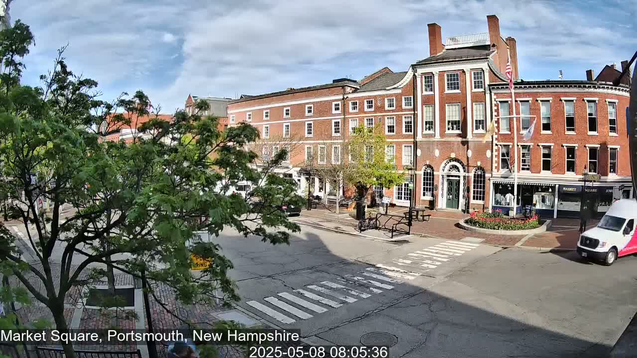 A mostly sunny day in a brick-lined town square, featuring several multi-story buildings, lush green trees, and a white van.