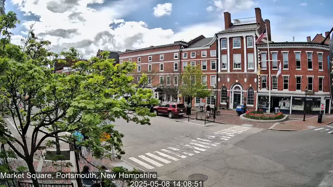 A partly cloudy day in a town square shows several brick buildings, green trees, a crosswalk, and a few parked cars.