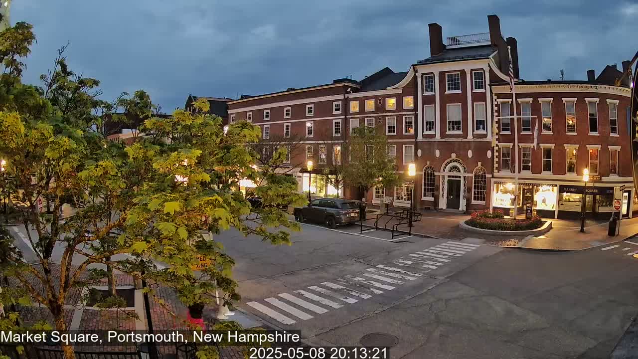 Under a twilight sky, a brick-lined town square is partially illuminated by streetlights, with a few cars and green trees present.