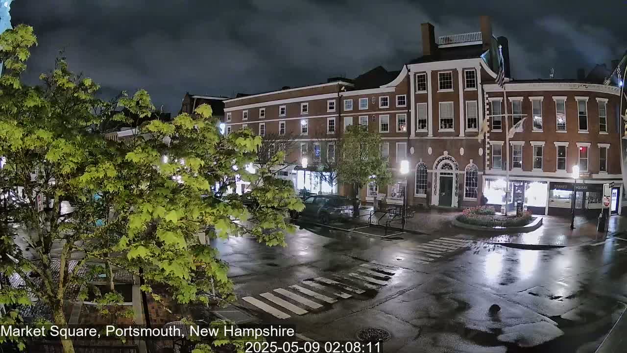 A nighttime view of a town square with brick buildings, wet streets, and lush green trees under a cloudy sky.