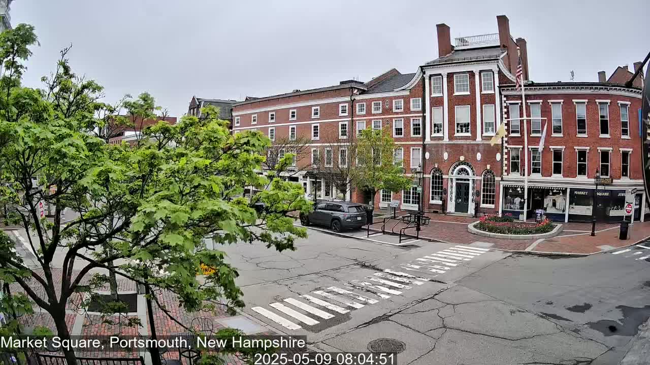 A partly cloudy day reveals a brick street with a crosswalk and several multi-story brick buildings, one of which has an American flag displayed in front, surrounded by lush green trees.