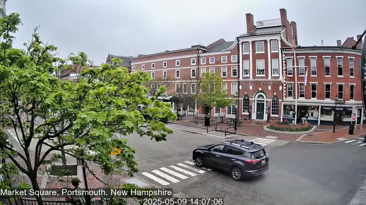 A dark-grey SUV drives across a crosswalk in a brick-lined town square under an overcast sky, past several multi-story brick buildings with lush green trees.