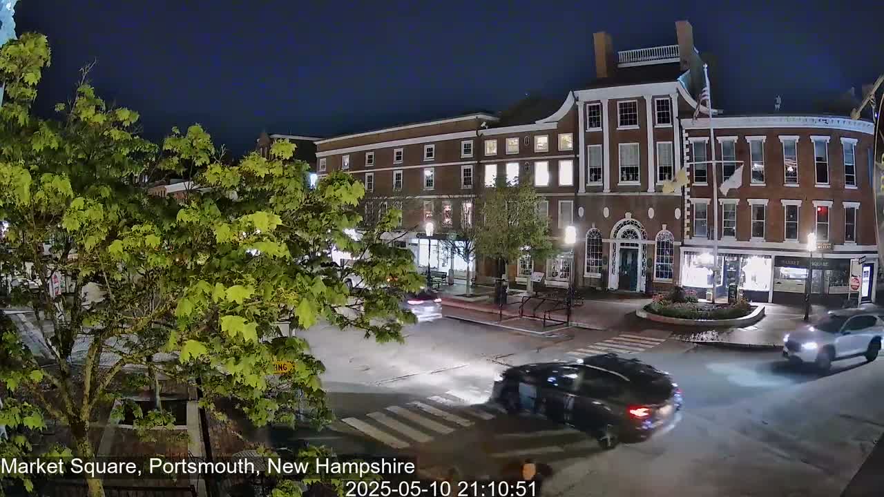 At night, several cars drive through a town square lined with brick buildings and illuminated storefronts, under a clear, dark sky.