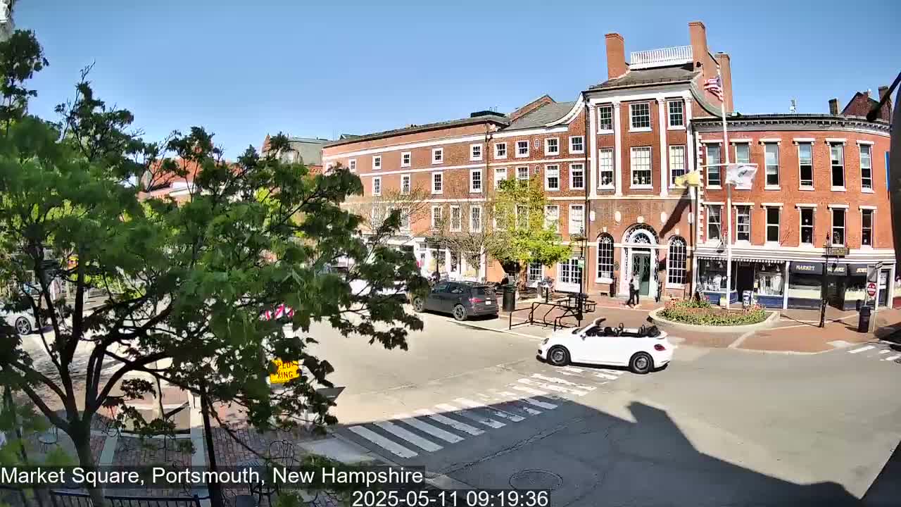 A sunny day reveals a town square with brick buildings, lush green trees, and a few cars, including a white convertible.