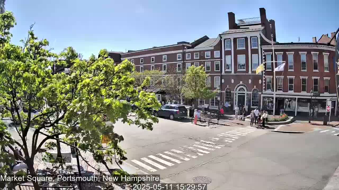 A sunny day reveals a town square with brick buildings, lush green trees, pedestrians crossing a street, and several cars parked nearby.