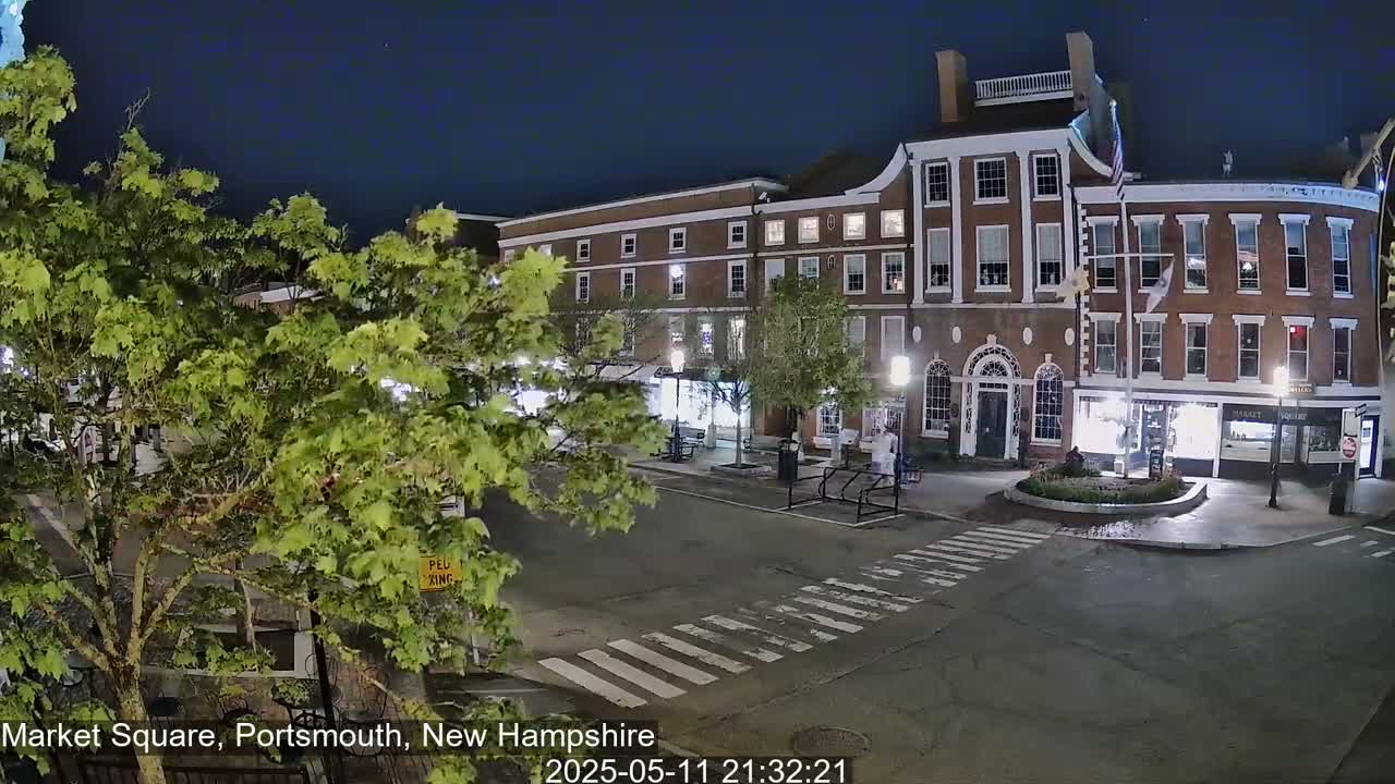 A nighttime view of a brick building and town square, with trees and a crosswalk, under a clear, dark sky.