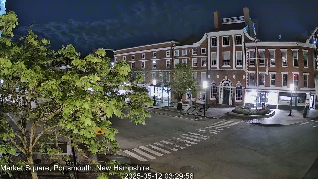 Under a partly cloudy night sky, a brick building and a tree stand in an empty town square.