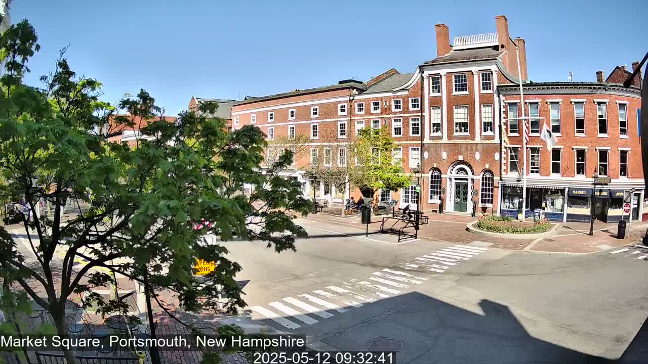 A sunny day reveals a brick-fronted town square with several buildings, trees, and pedestrians.