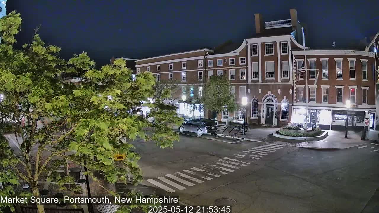 A nighttime view of a town square with brick buildings, streetlights, and a few cars, under a clear, dark sky.