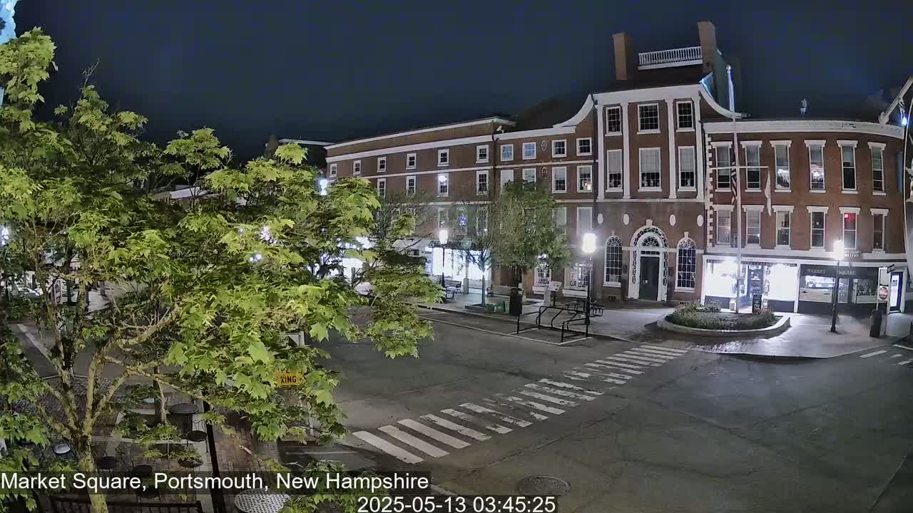 A mostly empty town square at night, under a dark sky, is lit by streetlights and shows a brick building with several storefronts.
