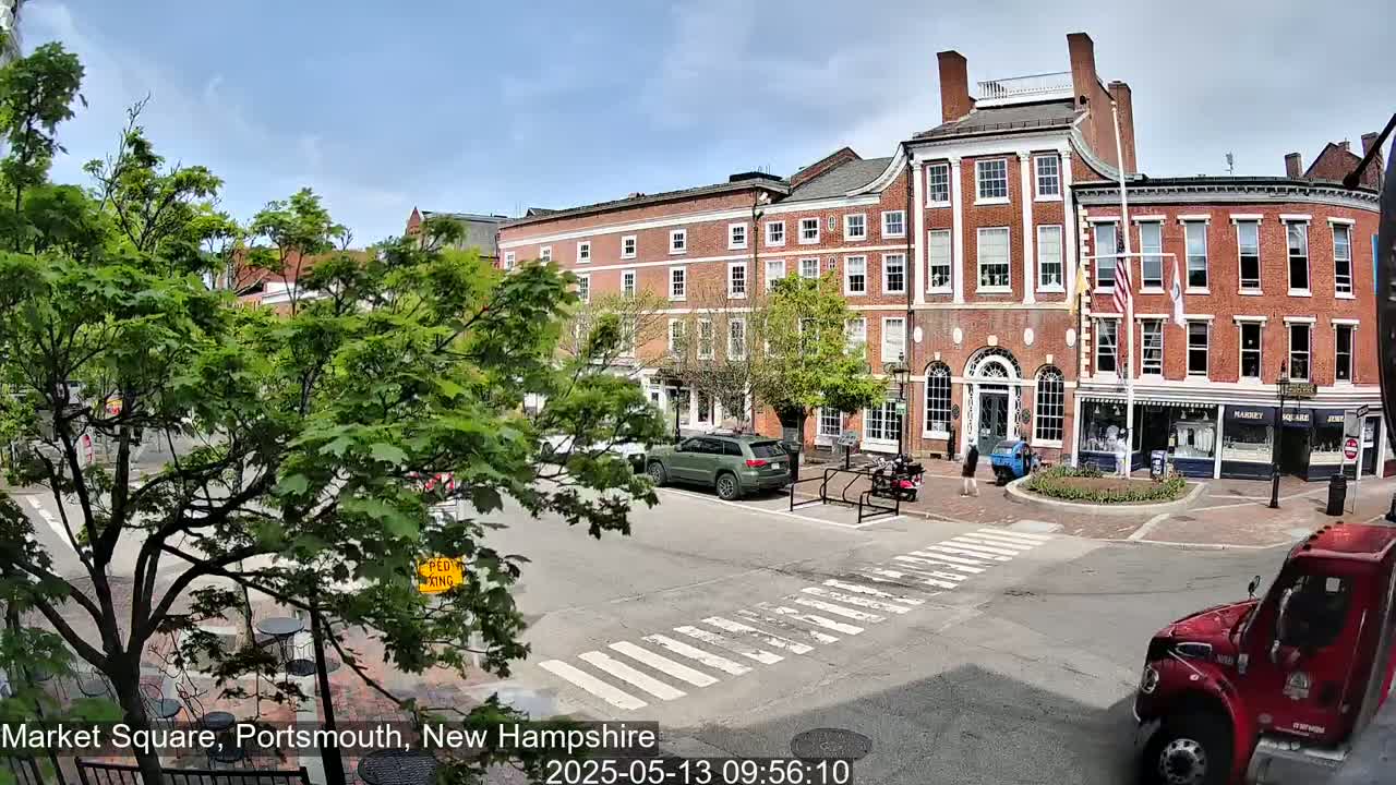 A partly sunny day shows a brick building in a town square with cars parked and pedestrians walking, and a red truck is visible in the bottom right corner.