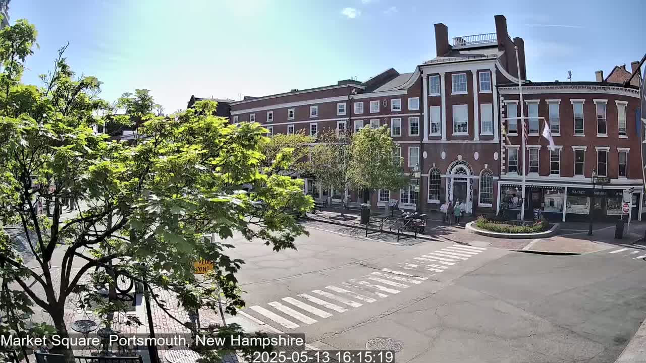 A sunny day shows a brick building in a town square with crosswalks and several leafy green trees.