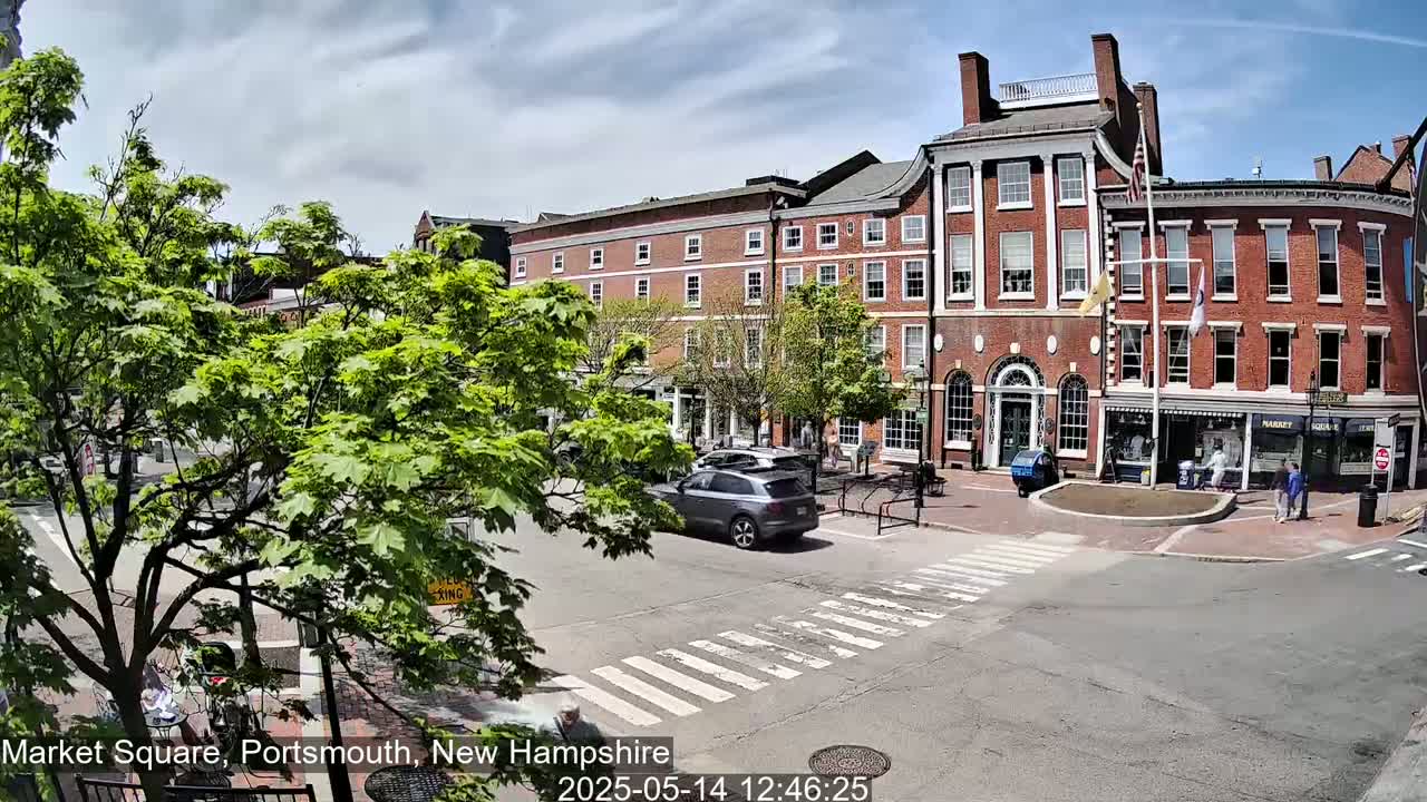 A sunny day in a town square shows a brick building and several cars, with pedestrians crossing a crosswalk near lush green trees.