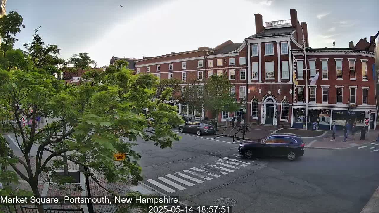 A partly cloudy evening scene shows a brick building in a town square, with cars and pedestrians at a crosswalk and lush green trees lining the street.