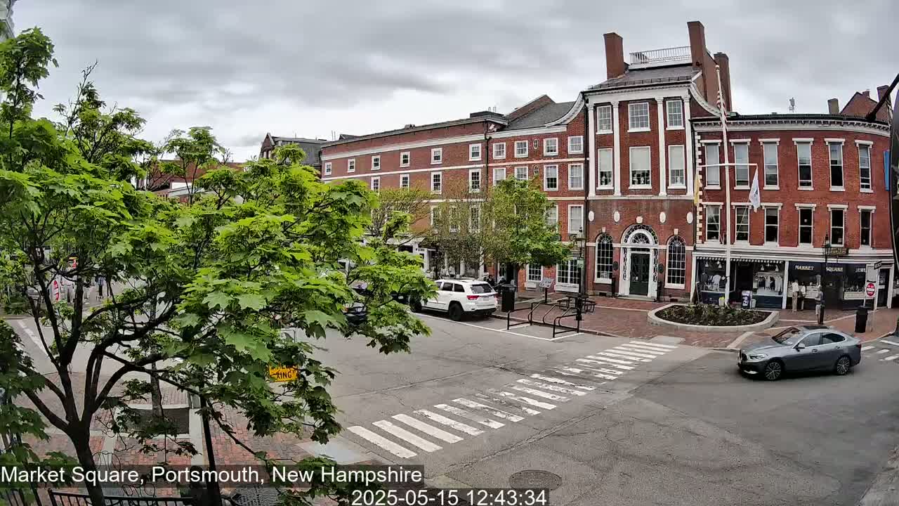 A street scene under a cloudy sky shows a brick building, several cars, and green trees in a town square.