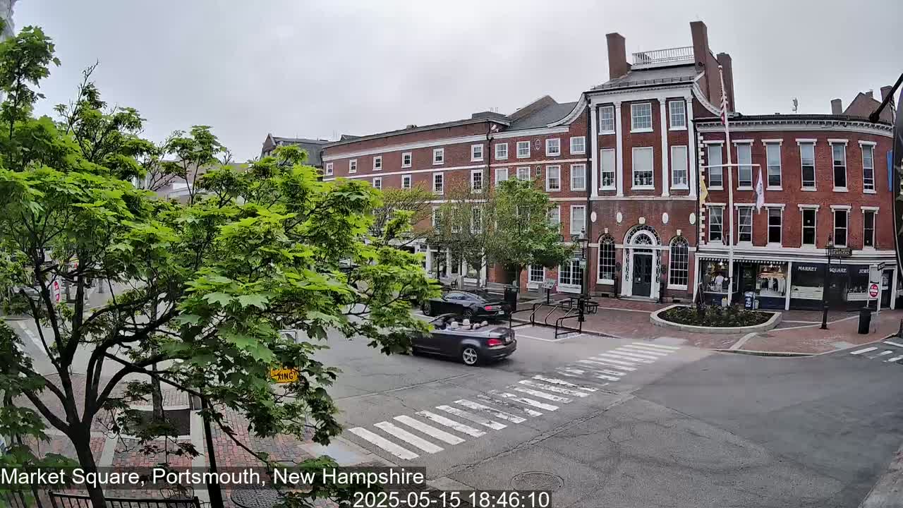 A street scene under a cloudy sky shows a brick building, several cars, and green trees in a town square.