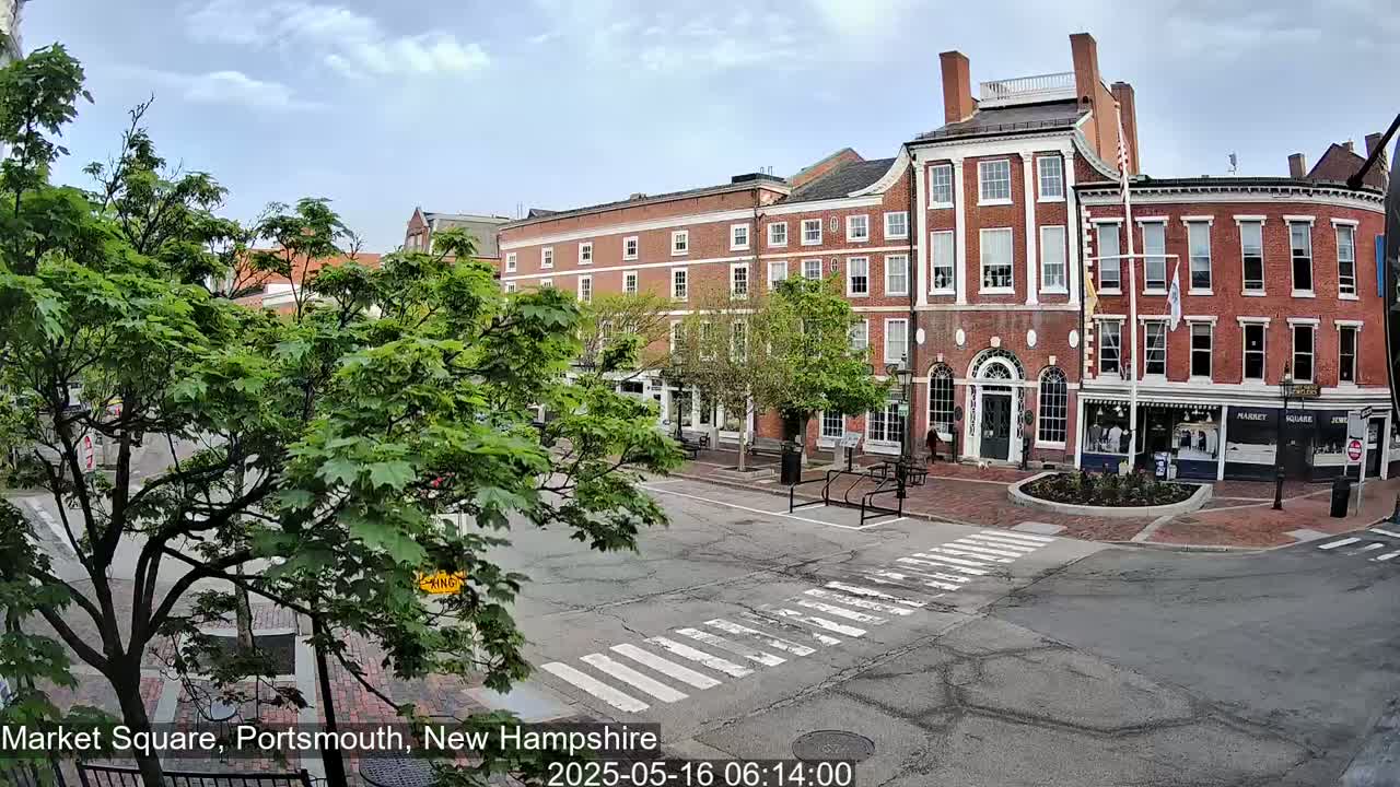 A partly cloudy day in a town square shows several brick buildings and lush green trees surrounding a paved pedestrian crossing.