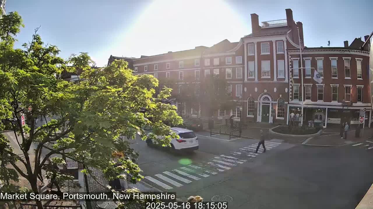 A sunny day shows a city square with brick buildings, pedestrians crossing a street, and a white SUV driving by.