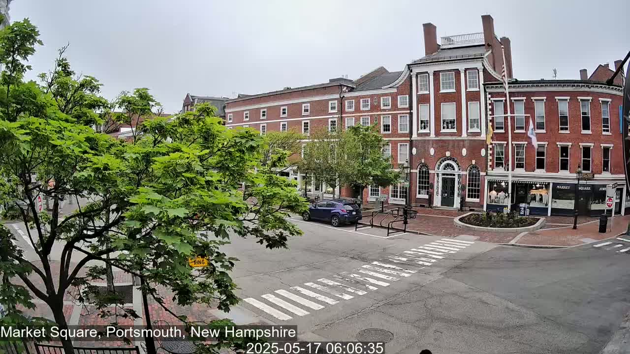 A mostly sunny view of a brick building in a town square, with a car parked across the street and lush green trees in the foreground.