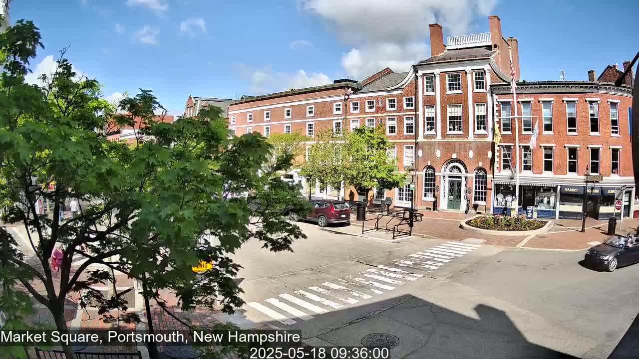 A sunny day in a brick-building-lined town square shows a few cars and pedestrians near a crosswalk, partially obscured by leafy green trees.