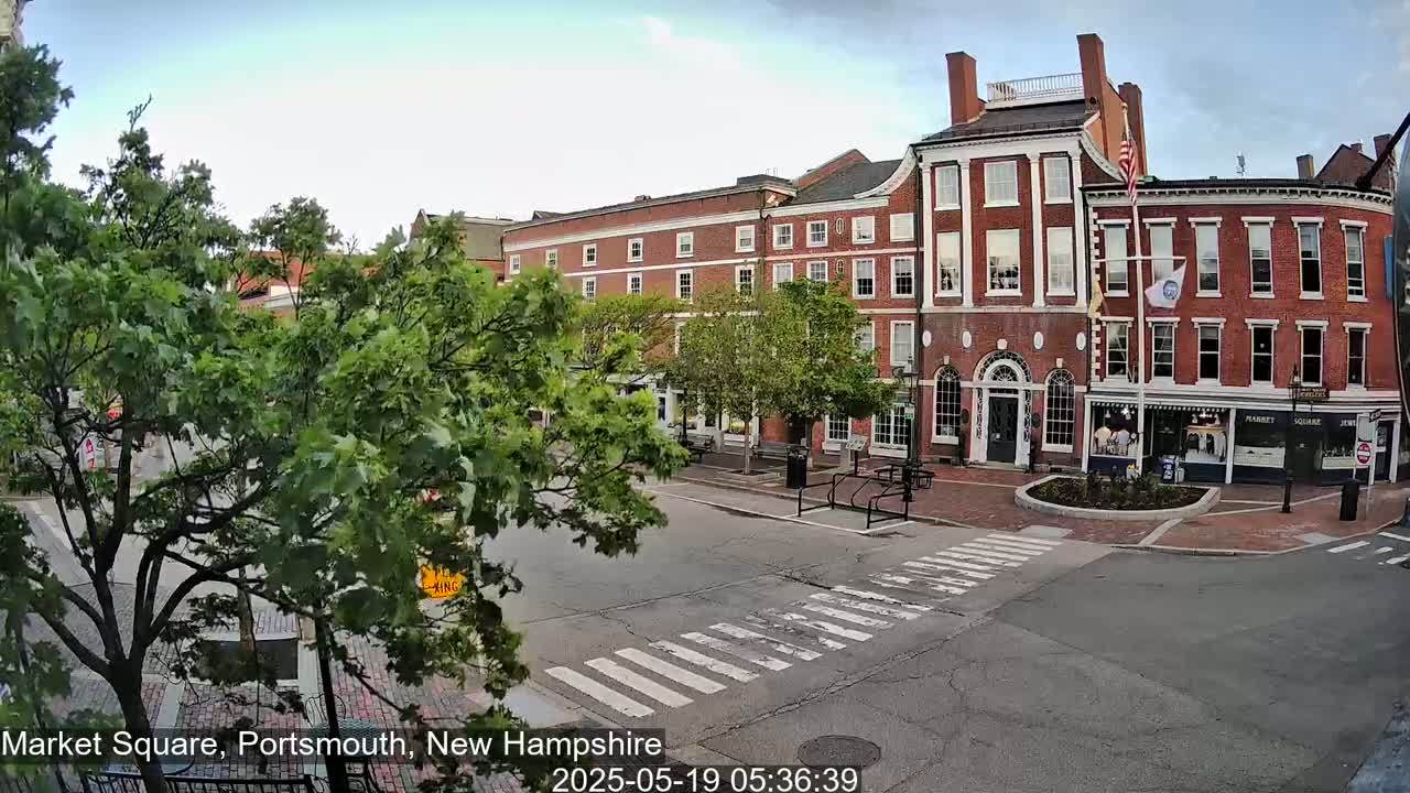 A partly cloudy day reveals a brick street scene with several buildings, lush green trees, and a pedestrian crossing.