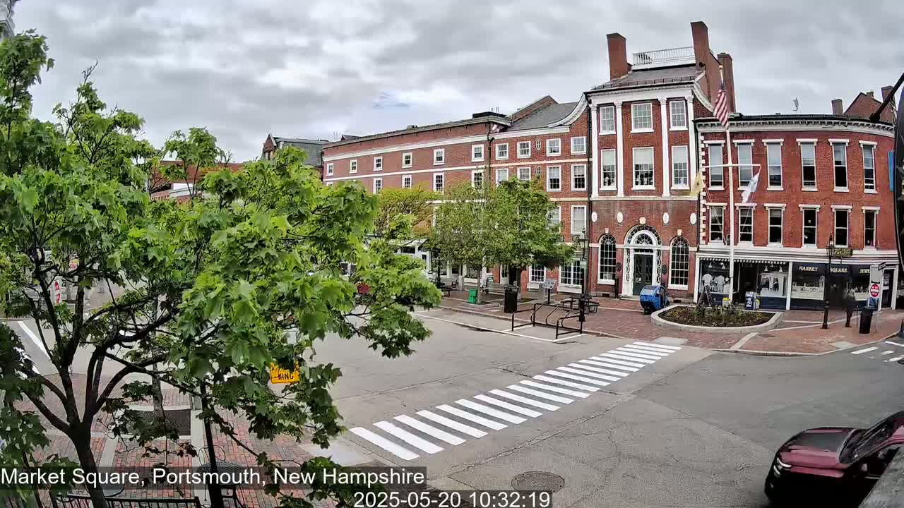 A brick building stands across a street with a crosswalk, under a cloudy sky, surrounded by lush green trees.