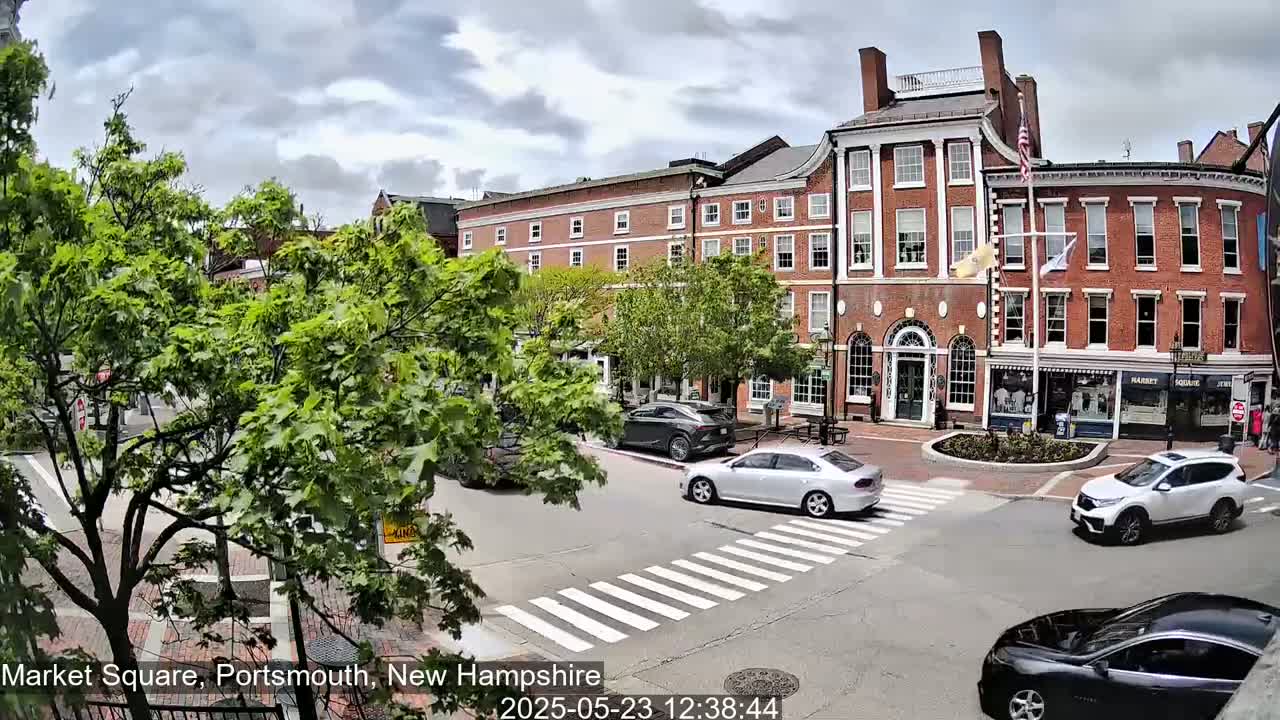 A partly cloudy day in a town square shows several cars at a crosswalk in front of brick buildings with lush green trees.
