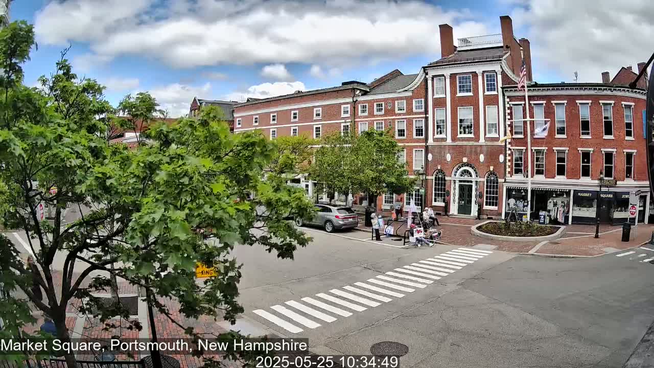 A partly sunny day in a town square shows a brick building with several shops on the ground floor and pedestrians crossing the street.