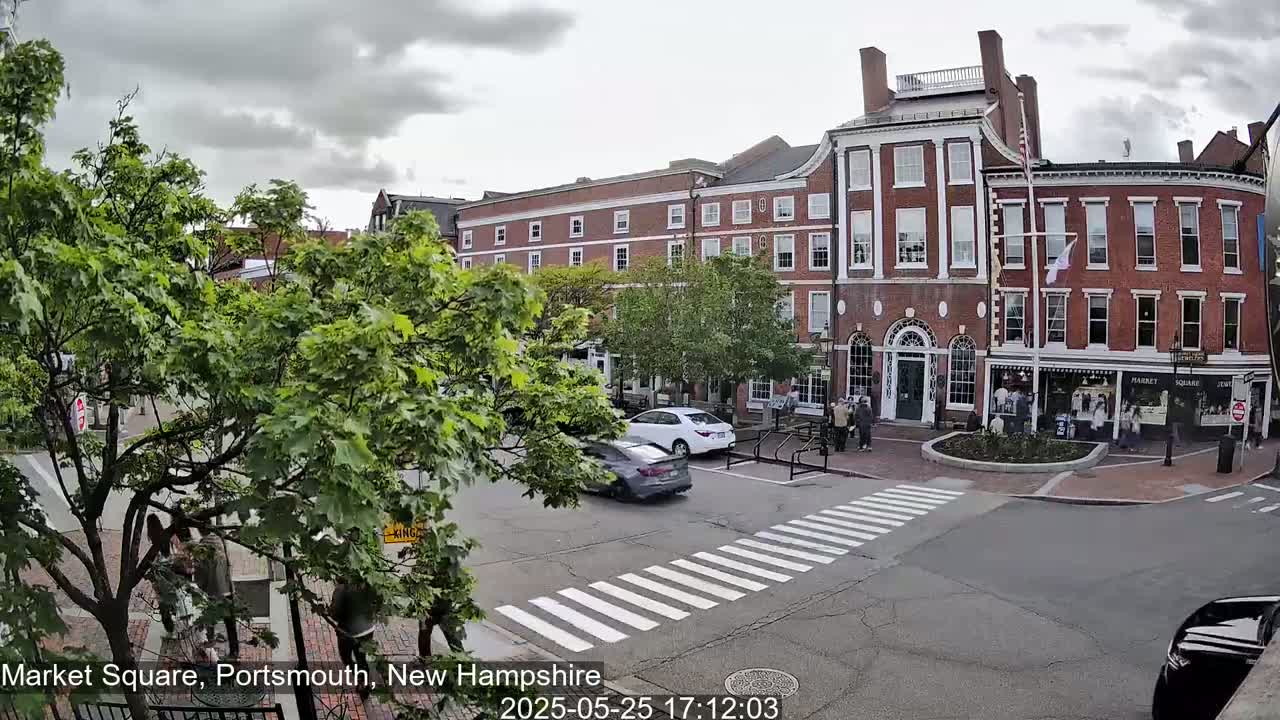 A view of a brick building and square in an overcast town, with several cars and pedestrians visible near a crosswalk.