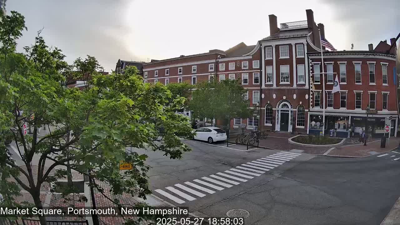 A mostly cloudy day reveals a brick building in a town square with several parked cars and pedestrians, surrounded by green trees.