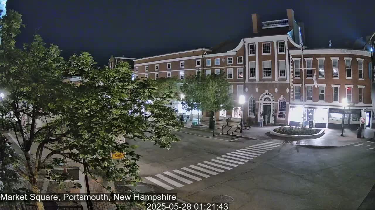 A nighttime view of a brick building in a town square, with trees and a crosswalk visible under a dark sky.