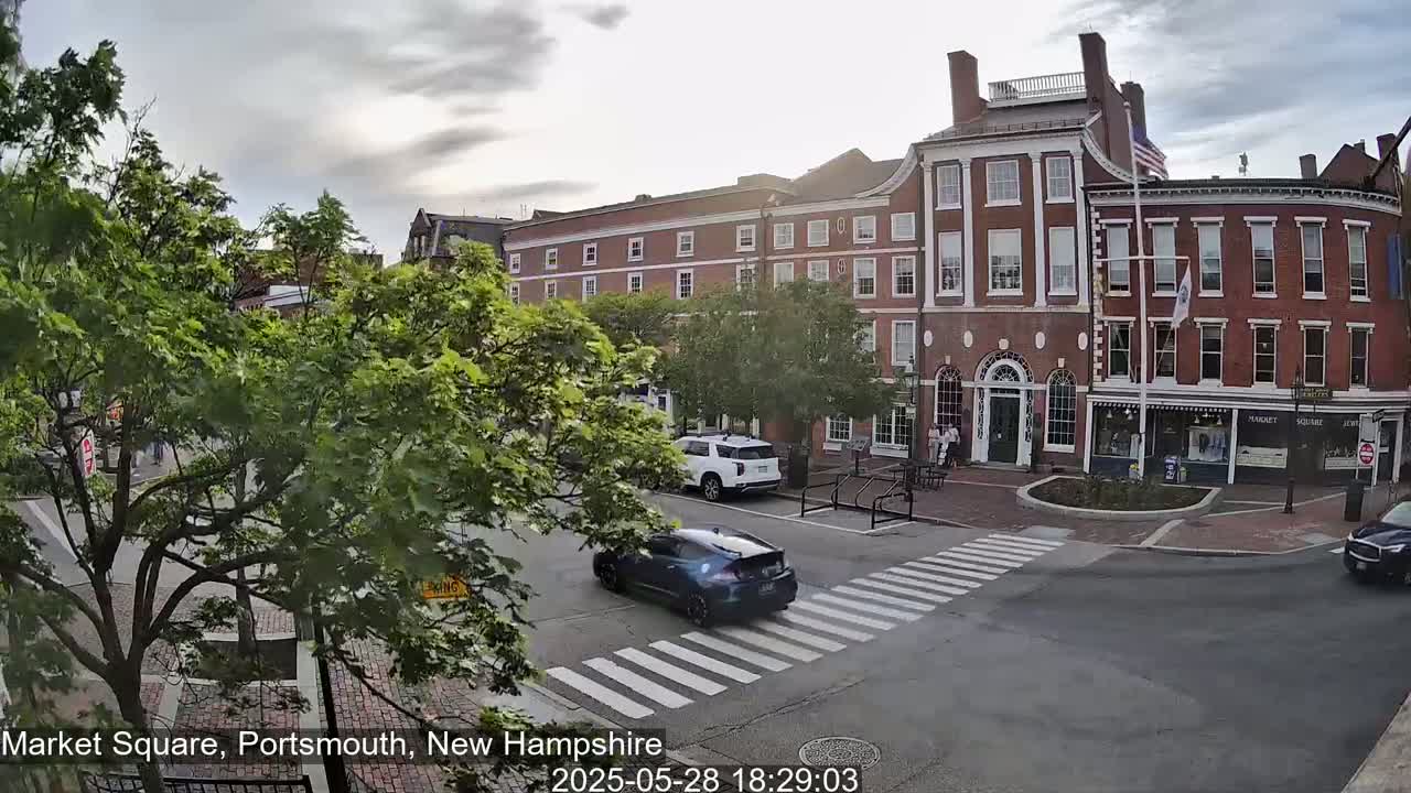 A brick building and a crosswalk are visible in a town square under partly cloudy skies, with several cars driving by and trees in the foreground.