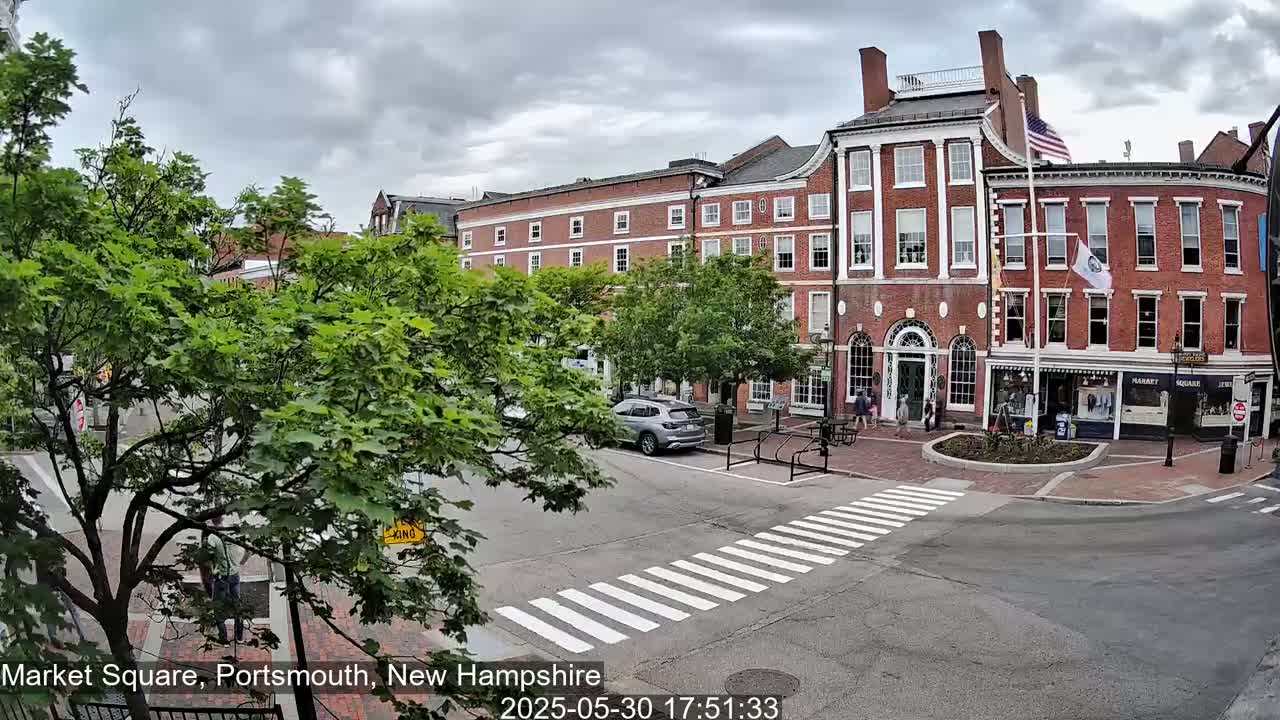 A street scene under a cloudy sky shows a brick building with shops on the ground floor and a crosswalk in the foreground.