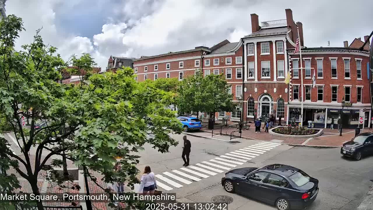 A partly cloudy day in a town square shows pedestrians crossing a street in front of brick buildings and parked cars.