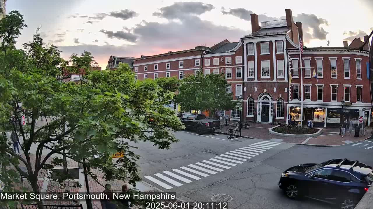 A brick building in a town square, with cars on the street and pedestrians crossing a crosswalk, under a partly cloudy evening sky.