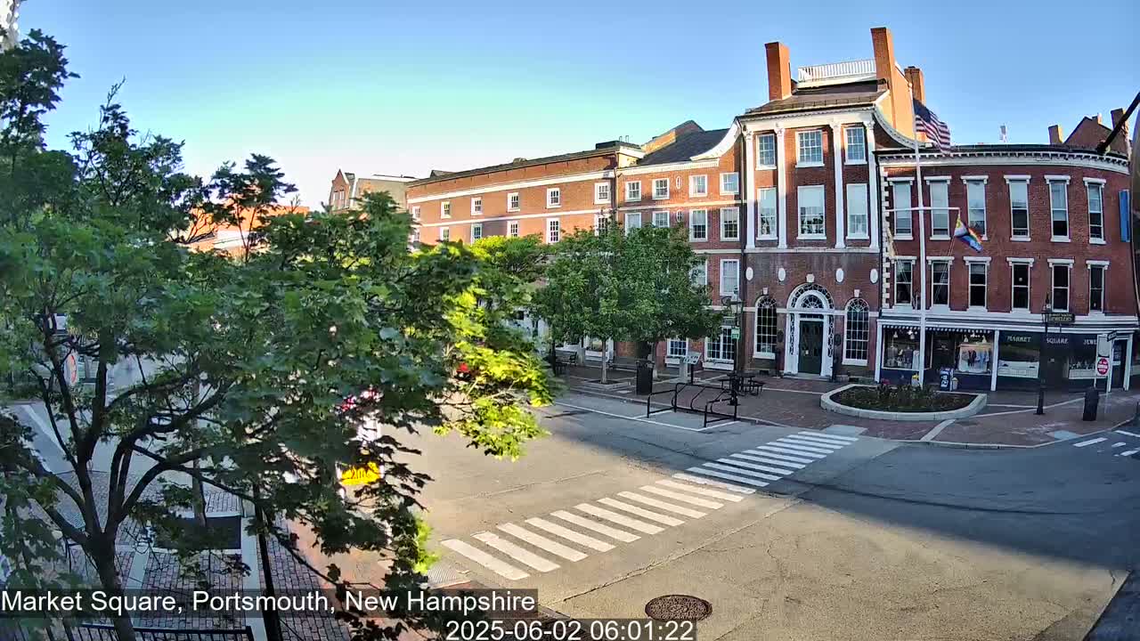 A sunny view of a brick building and a town square with a crosswalk, some cars, and lush green trees.