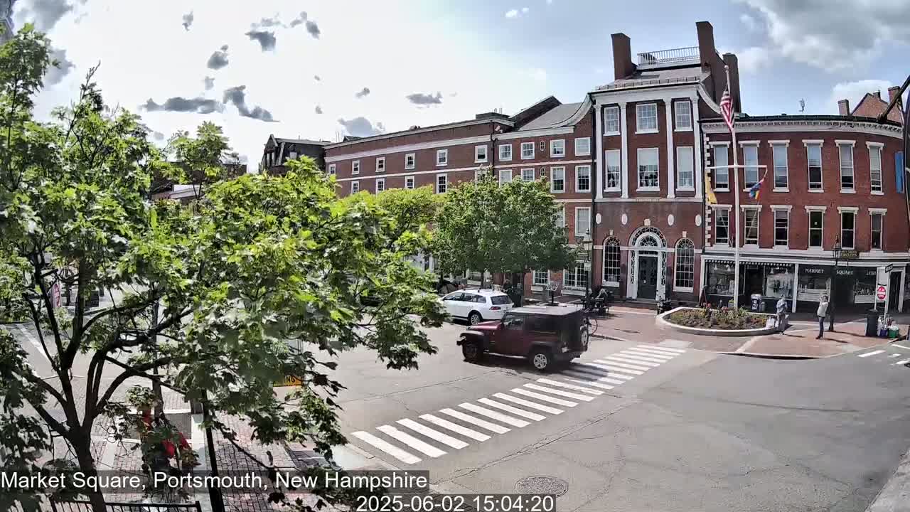 A partly cloudy day reveals a town square with several brick buildings, lush green trees, and a few vehicles including a red jeep driving through a crosswalk.