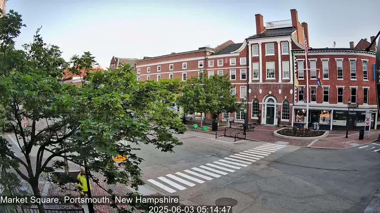 A brick building with many windows and a flagpole stands in a town square on a partly cloudy day.