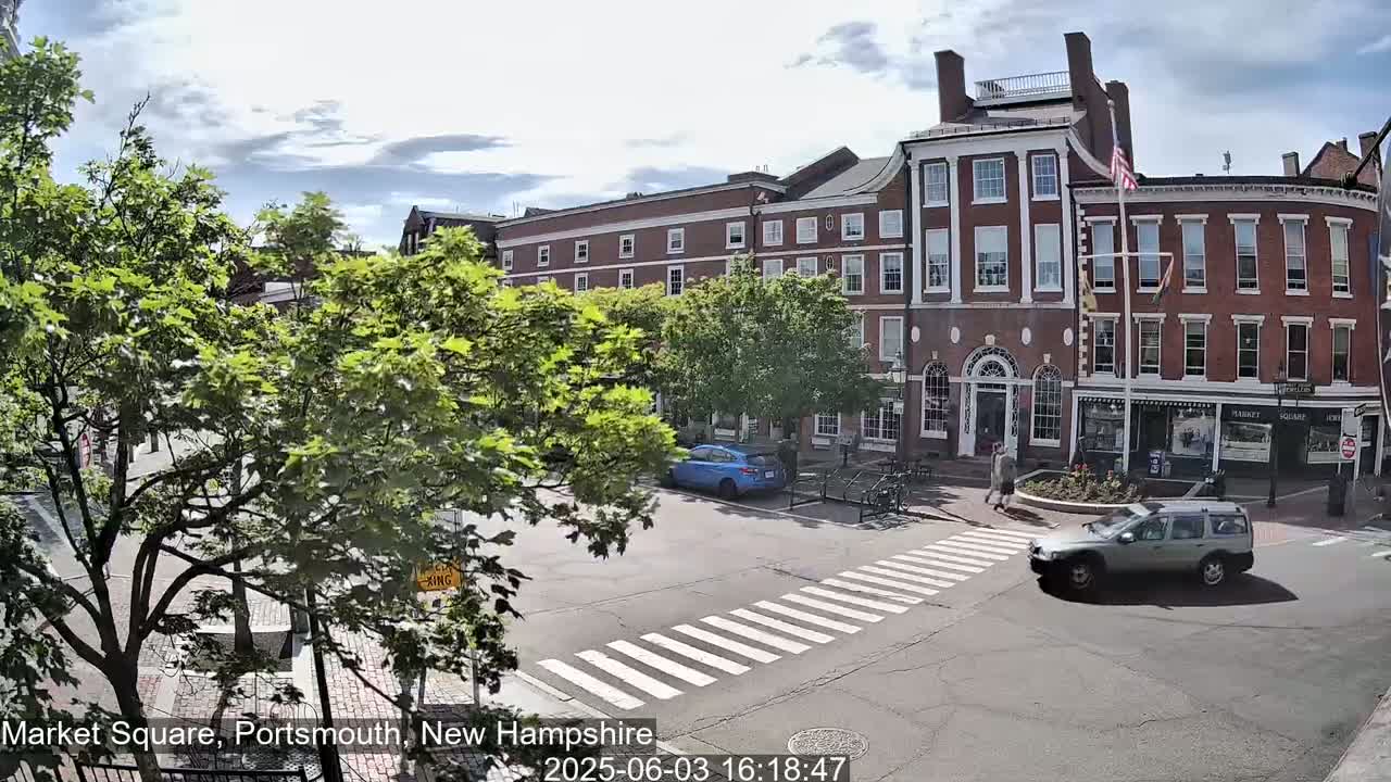 A partly sunny day reveals a town square with brick buildings, lush green trees, and a few cars and pedestrians at a crosswalk.