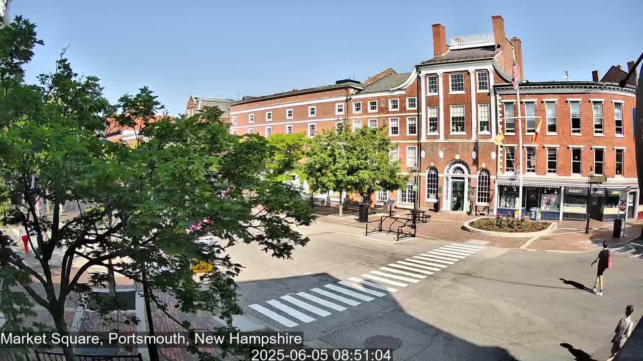 A sunny day reveals a brick building and shops surrounding a pedestrian crossing in a town square.