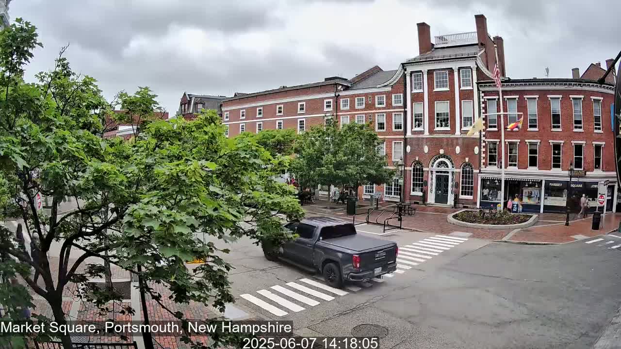 A gray pickup truck drives through a town square on an overcast day, passing in front of brick buildings with green trees lining the street.