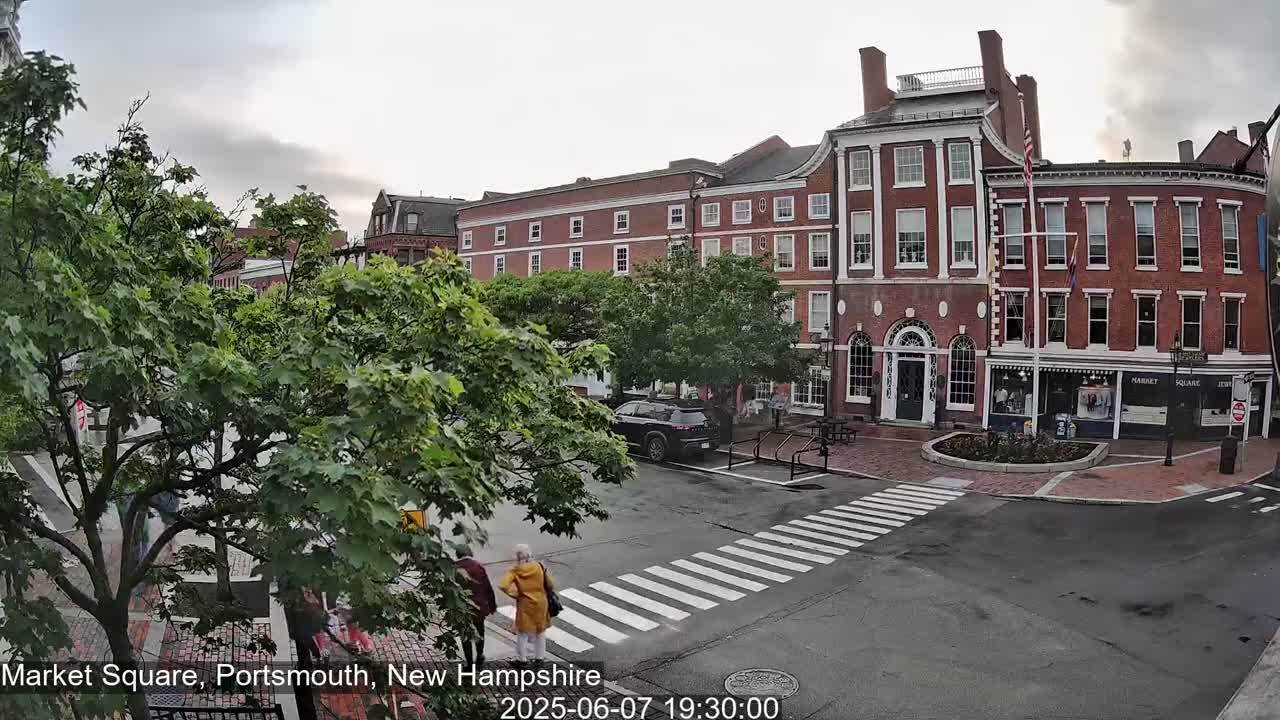 A partly cloudy day reveals a brick town square with pedestrians crossing a crosswalk in front of several multi-story buildings.