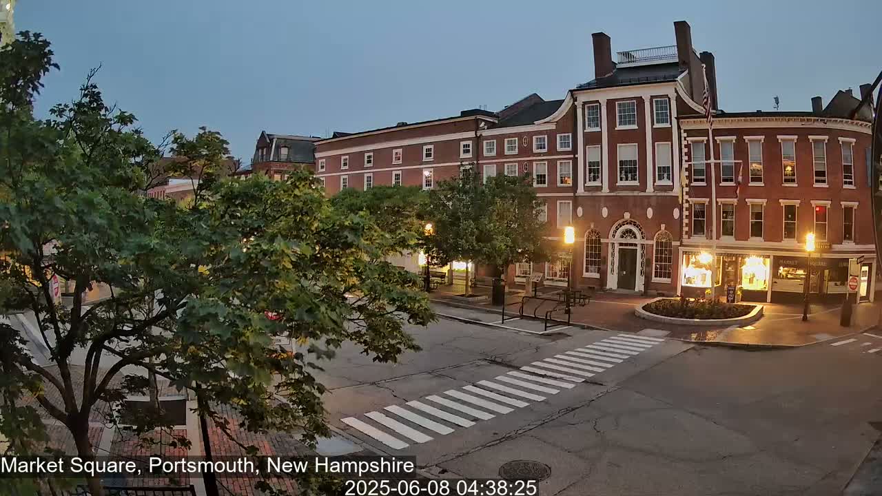 A twilight view of a town square with brick buildings, streetlights, and lush green trees under a clear, dusky sky.