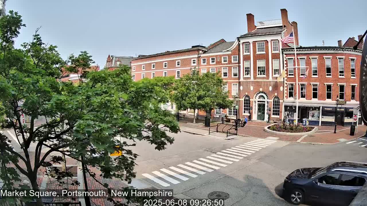 A sunny view of a town square with brick buildings, lush green trees, and a crosswalk, with a dark-colored SUV visible in the bottom right corner.